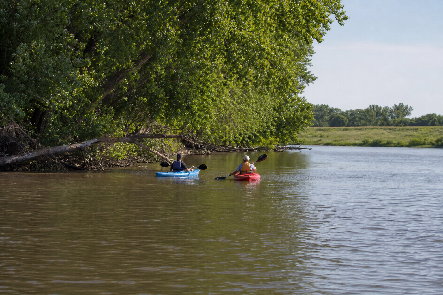 Two adults in life jackets paddle kayaks along a slow tree-lined river