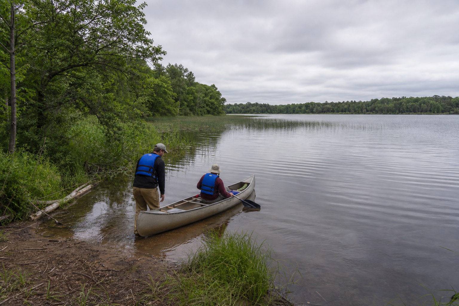 Two life-jacketed adults launch a canoe from a wooded natural lakeshore
