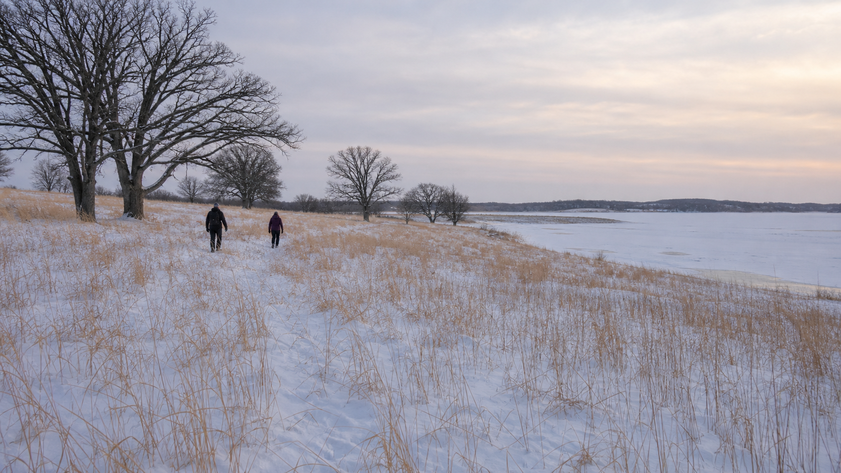 Two adults snowshoe on an illustrative prairie land trail beside a frozen lake