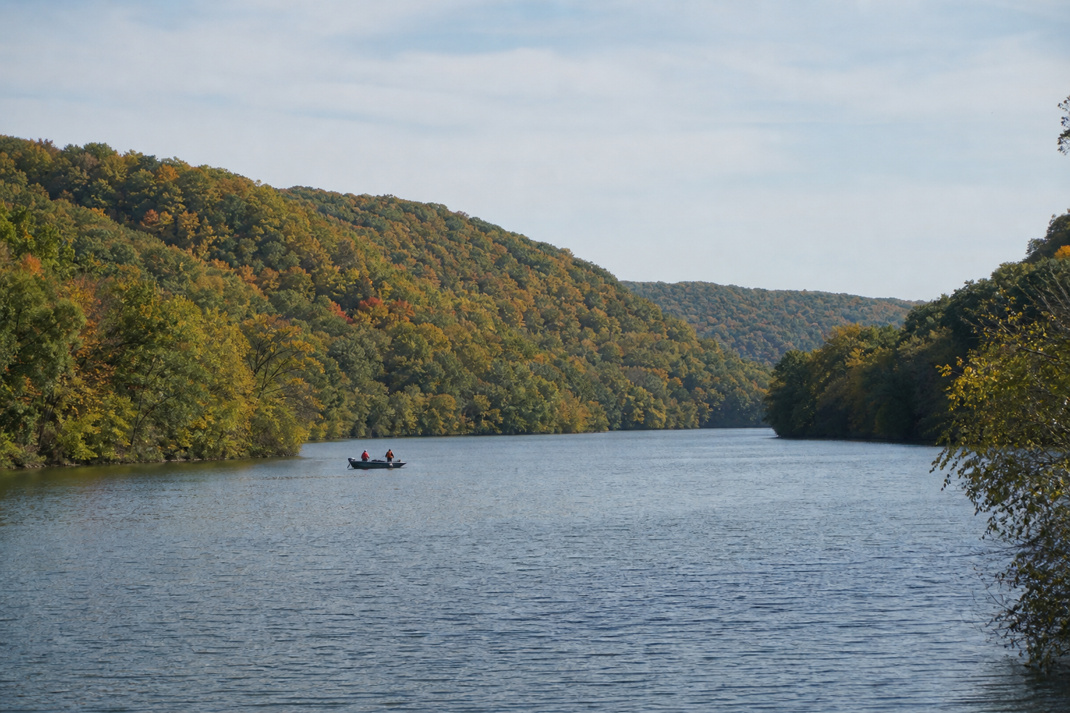 Two distant adults wearing life jackets fish from a small boat on an illustrative bluff-country river impoundment