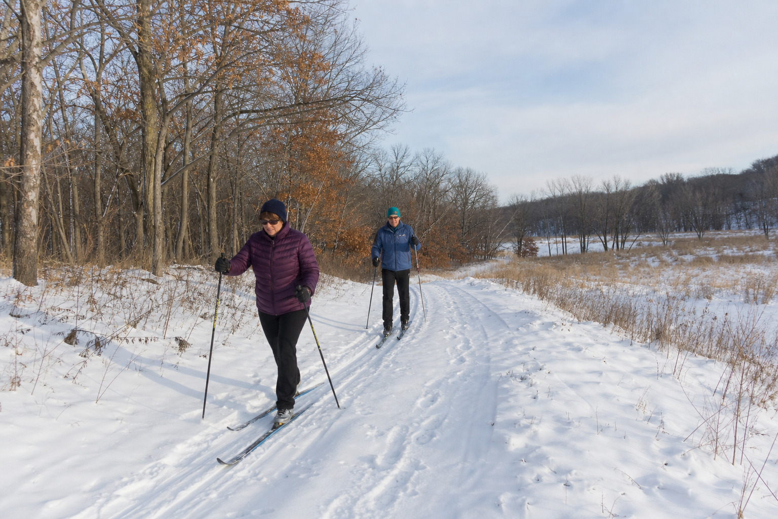 Two adults cross-country ski on a snow-covered land trail through Todd County hardwoods