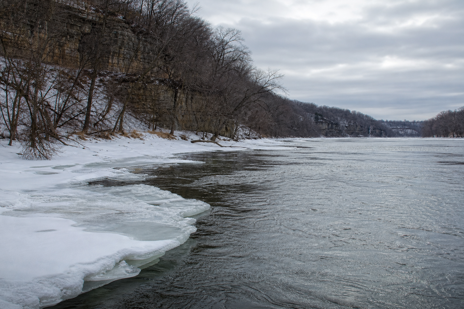 Illustrative snowy bluff-country riverbank beside open current and broken uneven ice.